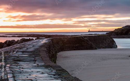 The stone pier at the harbour mouth of Cullercoats Bay on the coast of North Tyneside, England, UK. At sunrise with a colourful sky, taken at low tide.