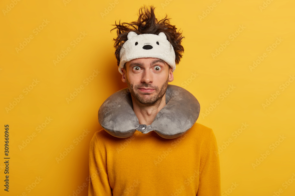 Φωτογραφία Funny unshaven man wears sleep mask, neck pillow, sleeps while traveling in car, has messy hairstyle, wears yellow jumper, poses indoor