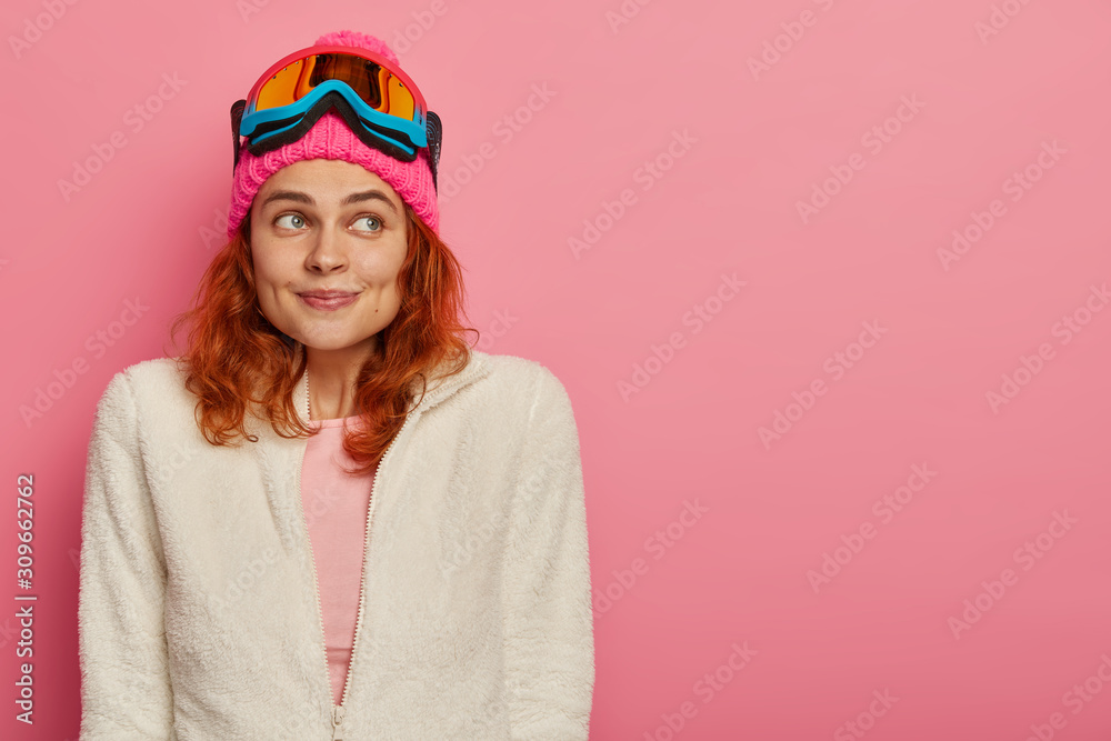 Satisfied young woman with ginger hair rides on snowboard during winter time, has dreamy expression, wears warm white jumper, thinks about next holiday trip, isolated on pink wall, copy space Satisfied young woman with ginger hair rides on snowboard during winter time, has dreamy expression, wears warm white jumper, thinks about next holiday trip, isolated on pink wall, copy space