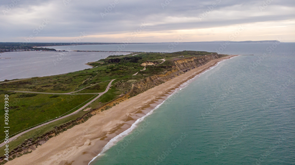 An aerial view of the Hengistbury Head with majestic sandy beach, crystal green water, grassy hill, huge cliff with the harbor and sea in the background undef a grey sky