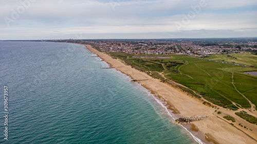 An aerial view of a sandy beach with groynes (breakwaters), crystal blue water, dunes, grassy field and city in the background under a grey sky © Dolwolfian