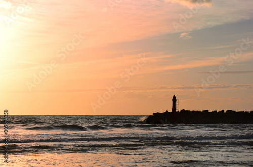 Dramatic sunset by the sea with a red sky reflecting on the sea and the waves behind the silhouette of a lighthouse on the coast © leopictures