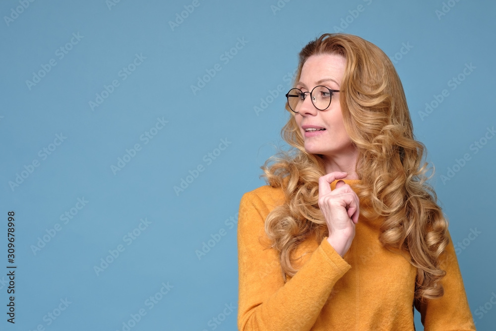 Mature caucasian happy woman with glasses and long blonde hair standing isolated over blue background looking aside.
