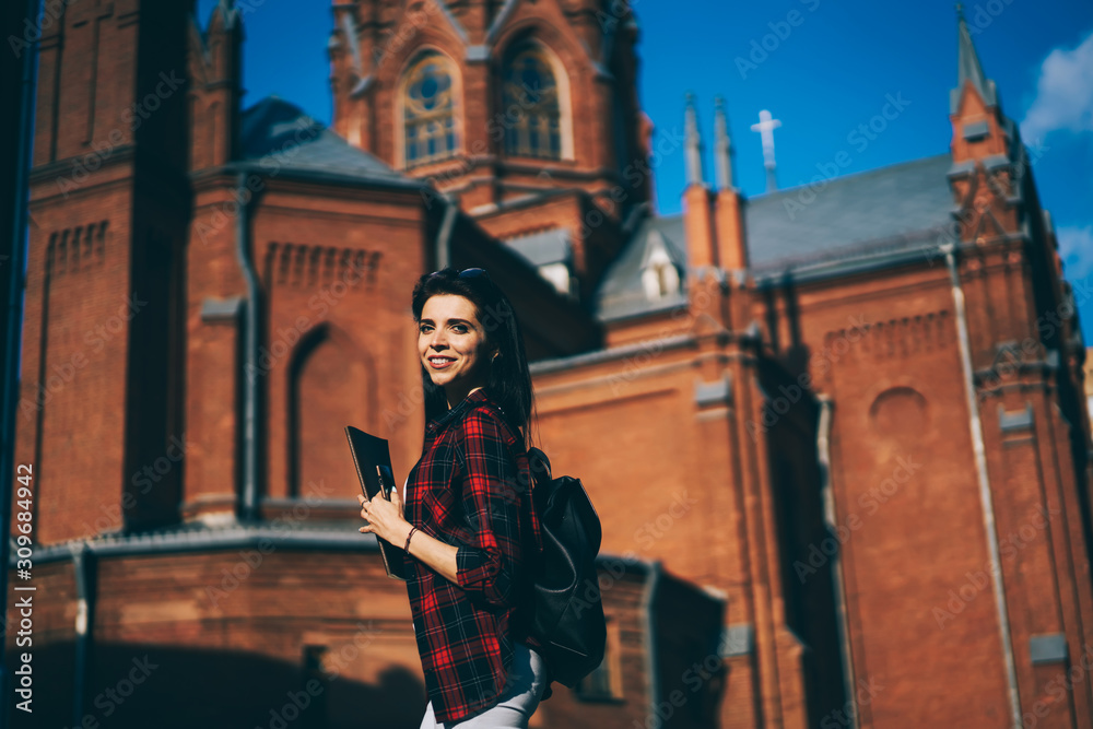 Successful smiling young hipster girl 20s walking at street and enjoying summer weather at square background, portrait of positive brunette woman strolling in downtown while looking at camera