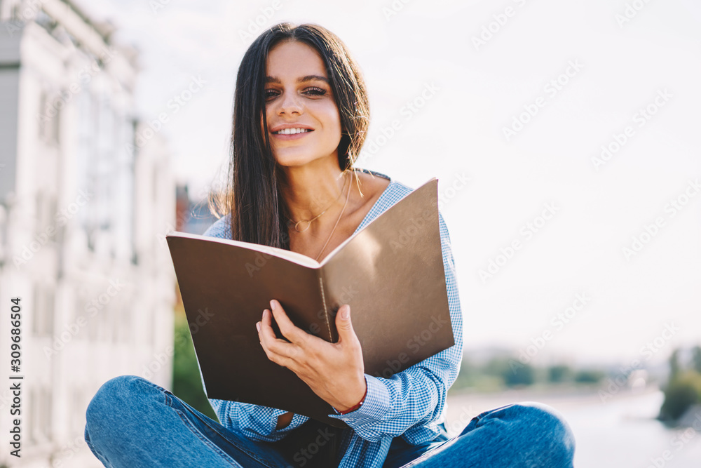 Obraz premium Adorable young student girl with brunette hair sitting and studying outdoors on bright summer day, portrait of attractive woman holding notebook doing homework looking at camera enjoying sunny weather