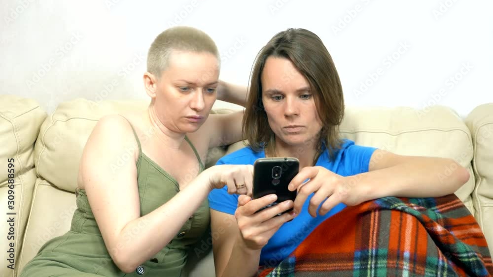 two women using a smartphone while sitting together on the sofa two women using a smartphone while sitting together on the sofa