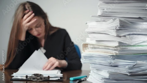 A pile of unfinished documents and files laying on desk in the office. Overworked female office accountant sitting in front of paperworks and holding her head with hands, tired worker having headache