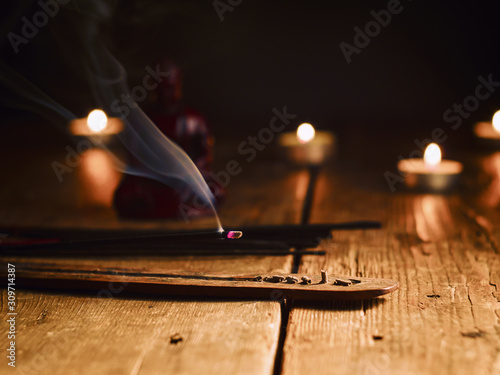 Smoking incense stick in the foreground. On background Small statue of Buddha with incense sticks and burning candles. Composition on old wooden table in dark room.