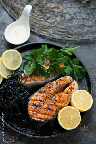 Fried salmon steaks with black spaghetti, lemon and parsley on a metal tray, vertical shot