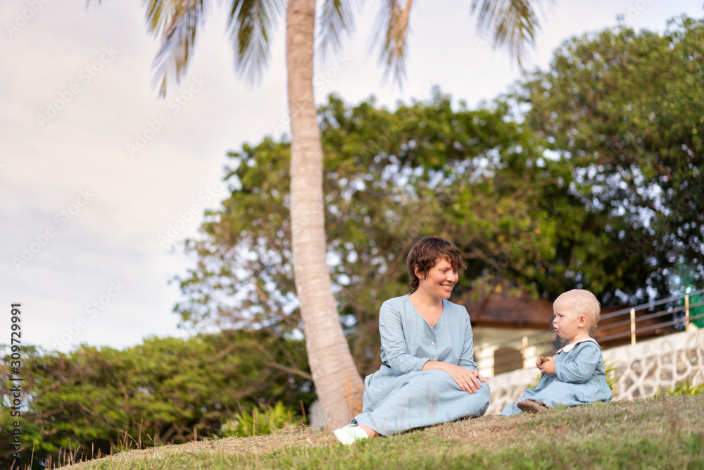 Beautiful woman mom sits under a palm tree with her daughter in blue dresses. Family holidays in the tropics. Beautiful woman mom sits under a palm tree with her daughter in blue dresses. Family holidays in the tropics.