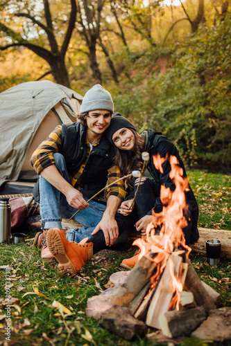 Young loving couple of tourists relaxing near the fire in the nature..Handsome man and beautiful woman are roasting marshmallows over the fire near tent in camping. Instagram stories picture.