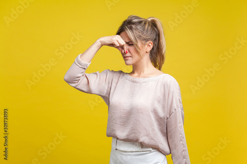 Unpleasant smell. Portrait of young dissatisfied woman with fair hair in casual beige blouse standing with closed eyes, pinching her nose with disgust. indoor studio shot isolated on yellow background