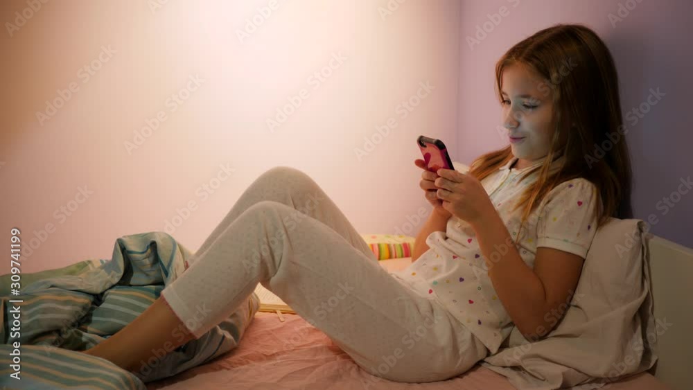 Cute kid girl in a bed text type smartphone messenger online pretending read a book when hears parent entering bedroom