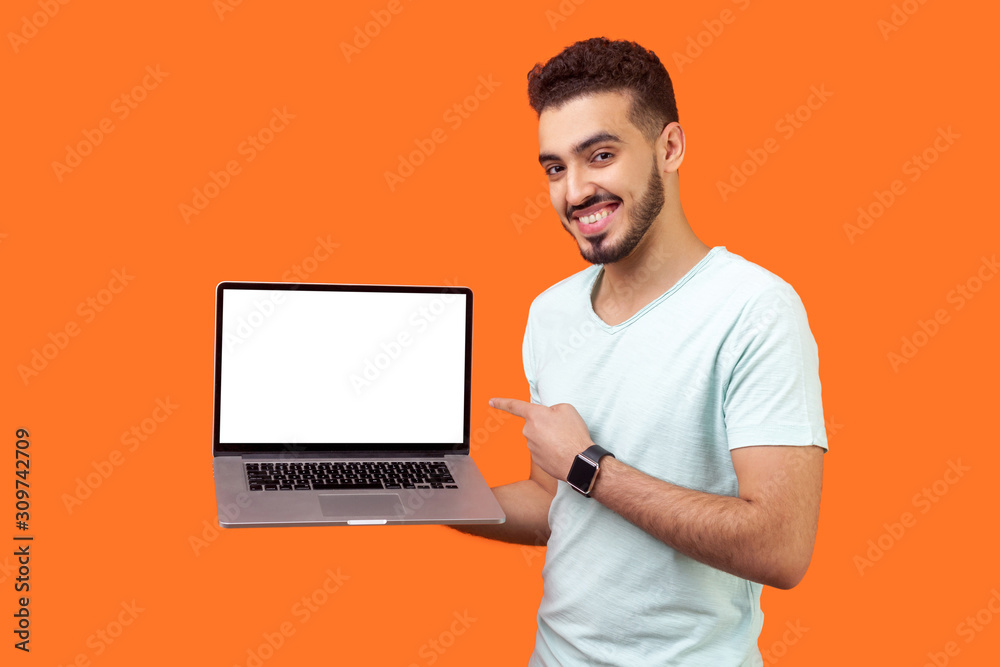 Portrait of positive joyful brunette man with beard in white t-shirt standing holding laptop with blank screen and smiling at camera, internet advertising. studio shot isolated on orange background Portrait of positive joyful brunette man with beard in white t-shirt standing holding laptop with blank screen and smiling at camera, internet advertising. studio shot isolated on orange background