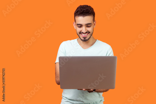 Portrait of cheerful young brunette man with beard in white t-shirt typing email on laptop and smiling, reading positive message or surfing the web. indoor studio shot isolated on orange background