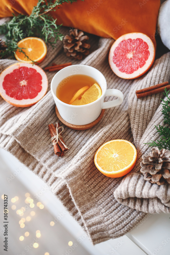 Christmas cozy winter home decor. New year still life: cup of citrus tea, oranges, grapefruits, cinnamon sticks, pine cones, fir branches, knitted sweater, glowing led garland lights. Christmas cozy winter home decor. New year still life: cup of citrus tea, oranges, grapefruits, cinnamon sticks, pine cones, fir branches, knitted sweater, glowing led garland lights.