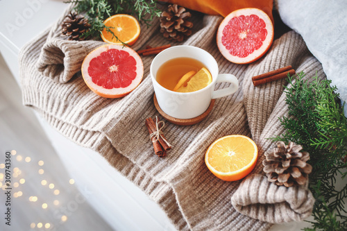 Christmas cozy winter home decor. New year still life: cup of citrus tea, oranges, grapefruits, cinnamon sticks, pine cones, fir branches, knitted sweater, glowing led garland lights.