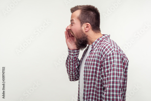 Side view of angry bearded man in casual shirt yelling loudly holding arm near mouth, shouting with aggressive expression, empty place for ad text, copy space. studio shot isolated on white background