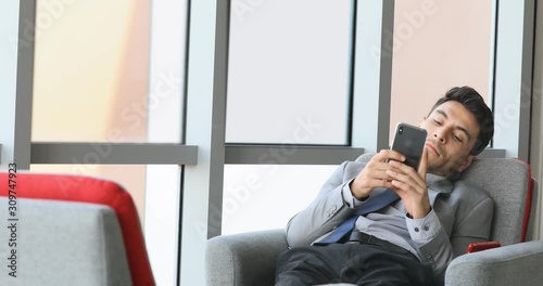 Handsome Caucasian businessman in grey suit sitting and using smartphone in modern office in his relaxing time.