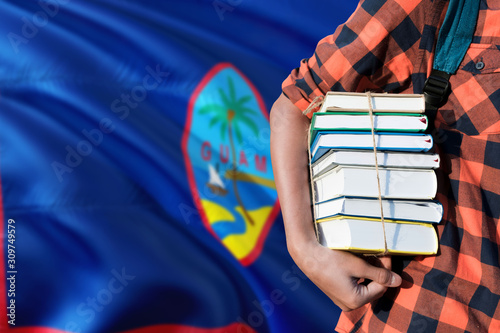 Guam national education concept. Close up of teenage student holding books under his arm with country flag background. © sezerozger