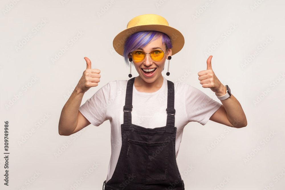 Like, well done! Portrait of stylish wonderful hipster girl with violet short hair in sunglasses and hat showing thumbs up gesture, success sign and smiling. isolated on white background, studio shot Like, well done! Portrait of stylish wonderful hipster girl with violet short hair in sunglasses and hat showing thumbs up gesture, success sign and smiling. isolated on white background, studio shot