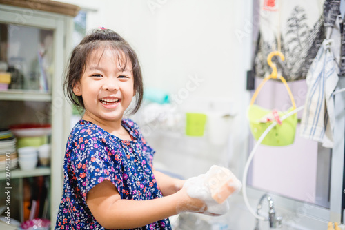 Obraz na plátně Happy smiley laughing face little cute asian girl washing dishes in kitchen for help her mom