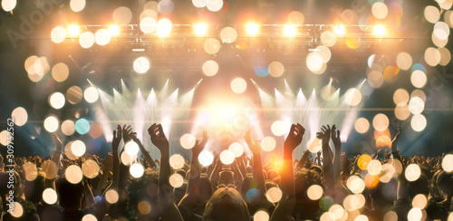 Photo of a concert hall with people silhouettes clapping in front of a big stage lit by spotlights. Shot is taken from concert crowd point of view, lens flare is visible. photo