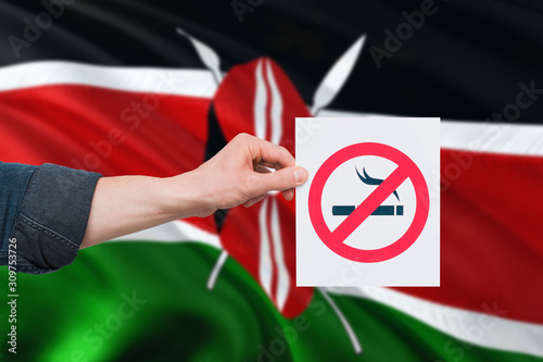 Kenya health concept. Hand holding paper with no smoking sign over national waving flag. Quit smoke theme.
