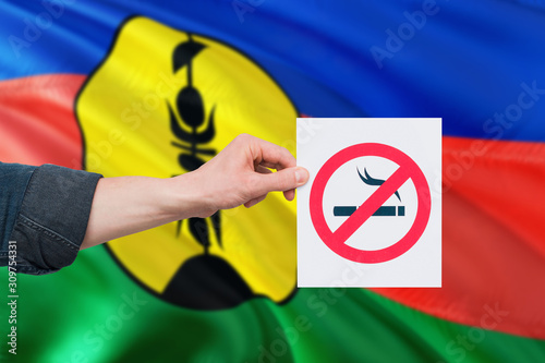 New Caledonia health concept. Hand holding paper with no smoking sign over national waving flag. Quit smoke theme.