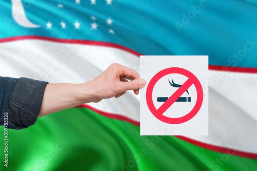 Uzbekistan health concept. Hand holding paper with no smoking sign over national waving flag. Quit smoke theme.