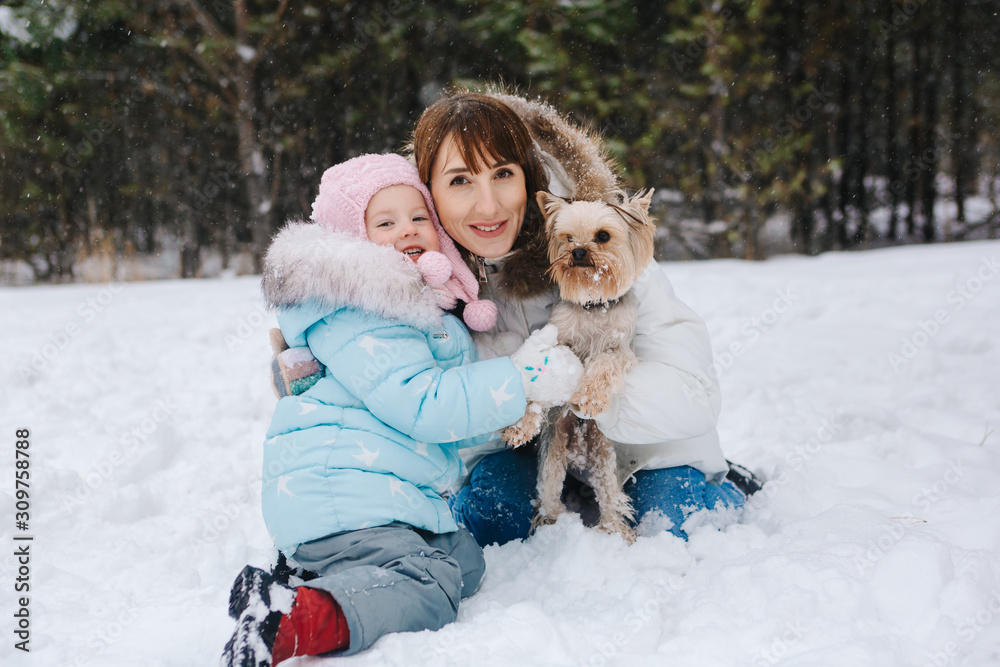 Obraz premium On a winter day, a woman and a girl are sitting in the snow against the background of a forest, hugging and holding a Yorkshire Terrier dog in their arms and looking at the camera.