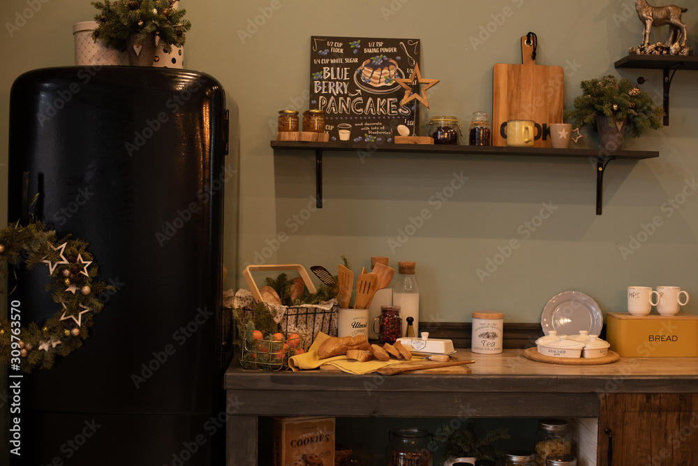 Christmas wreath and fir tree branches in vases and cups all around the modern kitchen. Modern kitchen with black fridge decorated for Christmas and New Year. Christmas wreath and fir tree branches in vases and cups all around the modern kitchen. Modern kitchen with black fridge decorated for Christmas and New Year.