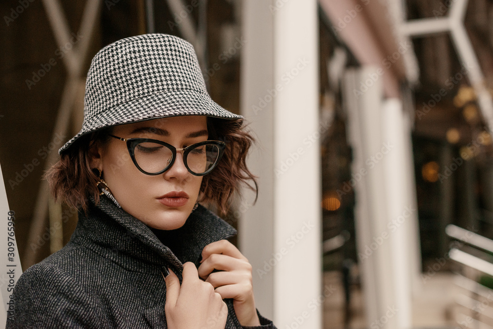 Outdoor close up fashion portrait of young elegant woman wearing trendy checkered bucket hat, stylish cat eye glasses, coat, posing in street of European city. Copy, empty space for text Outdoor close up fashion portrait of young elegant woman wearing trendy checkered bucket hat, stylish cat eye glasses, coat, posing in street of European city. Copy, empty space for text