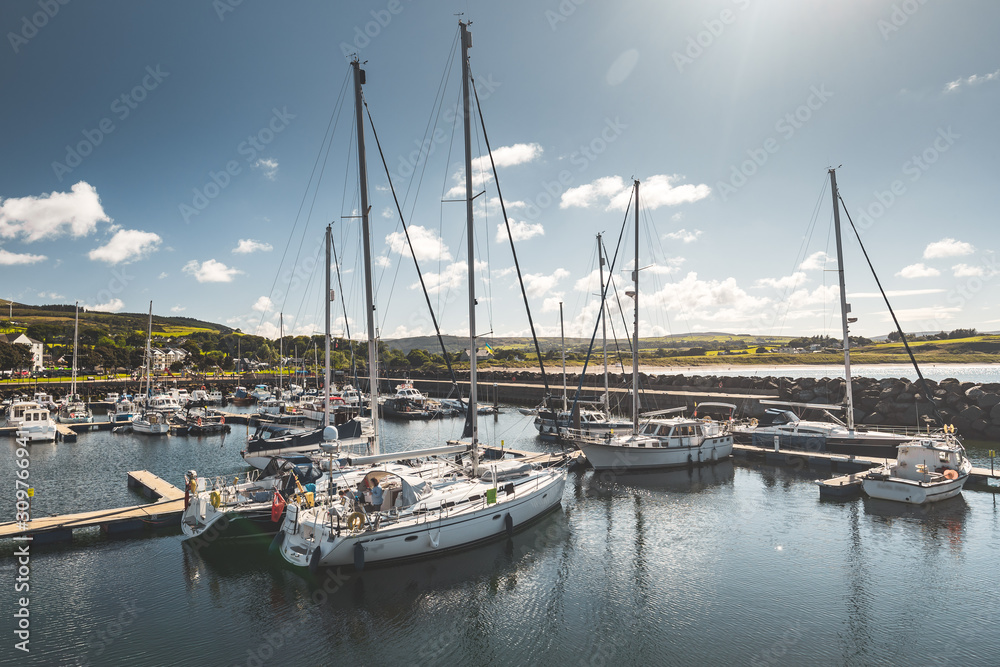 Numerous Yachts at Northern Ireland Club Pier. Sunny Sky with Clouds, Meadow and Green Hills on Background. Elegant Small Touristic Ships at Bay. Reflection in Water Surface. Luxury Lifestyle