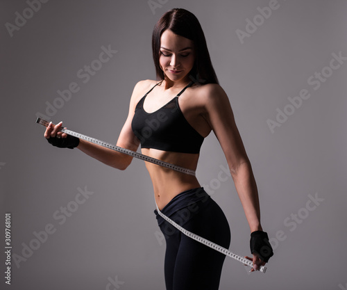 sporty woman with muscular body measuring her waistline with measure tape over gray