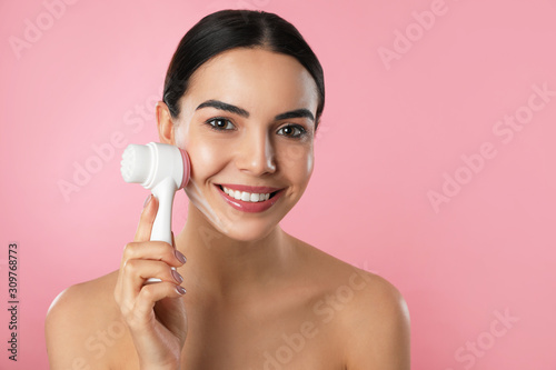 Young woman using facial cleansing brush on pink background. Washing accessory