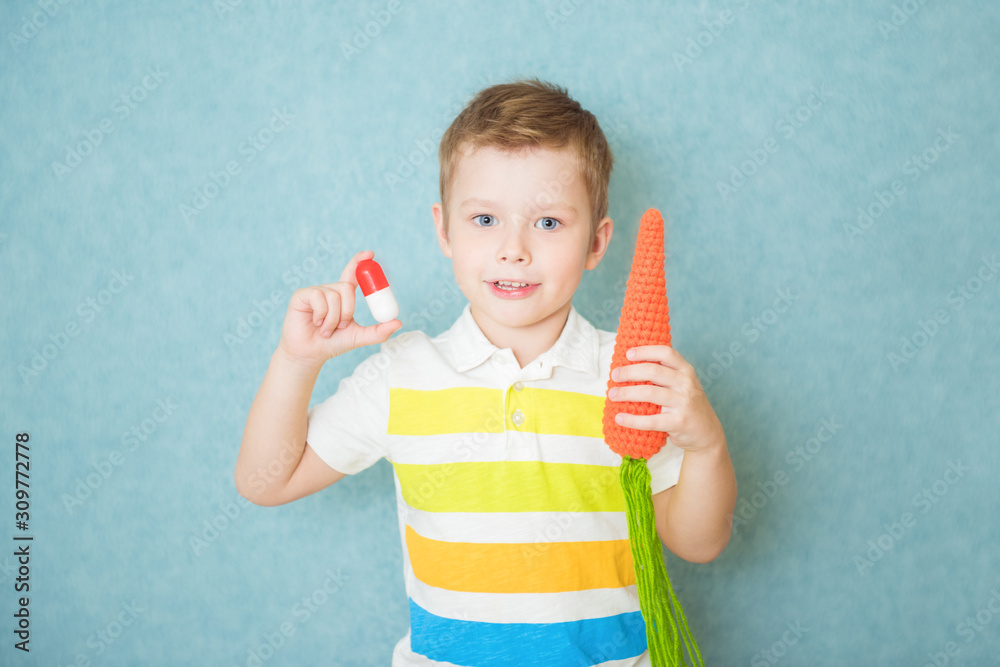 Smiling boy holding a capsule of vitamin and knit a carrot on blue background Smiling boy holding a capsule of vitamin and knit a carrot on blue background