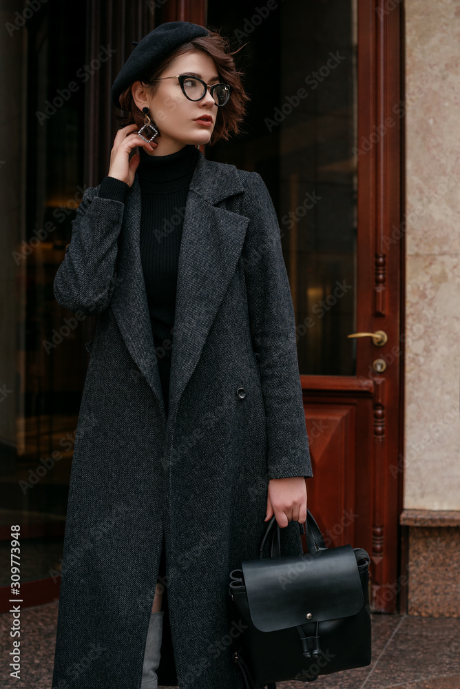 Outdoor fashion portrait of young elegant woman wearing total black outfit:  classic coat, turtleneck dress, glasses, beret, holding leather backpack,  posing in street of city Stock Photo | Adobe Stock