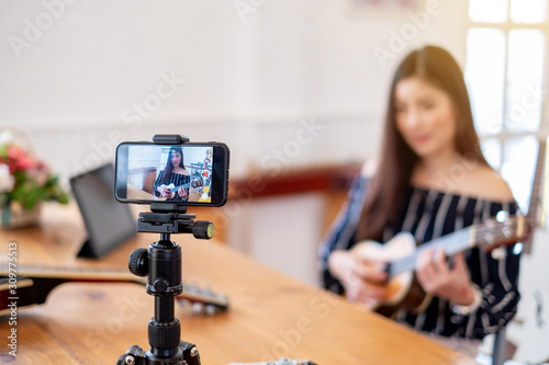Asian woman blogger playing acoustic guitar recording video clip by smartphone and camera at home.