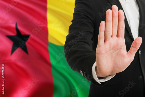 Guinea Bissau rejection concept. Elegant businessman is showing stop sign with hand on national flag background.