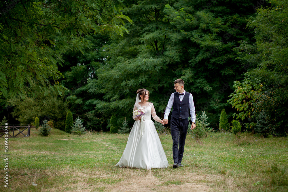 Happy bride and groom holding hands and walking in garden wedding day. Back view of charming stylish newlyweds holding hands while walking in park forest, happy marriage moments