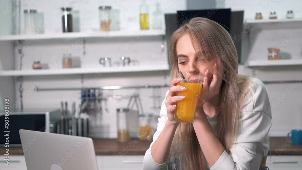 Young blonde woman drinking fresh juice at the kitchen. Working with notebook computer. Handheld shot. Slowmotion. Young blonde woman drinking fresh juice at the kitchen. Working with notebook computer. Handheld shot. Slowmotion.