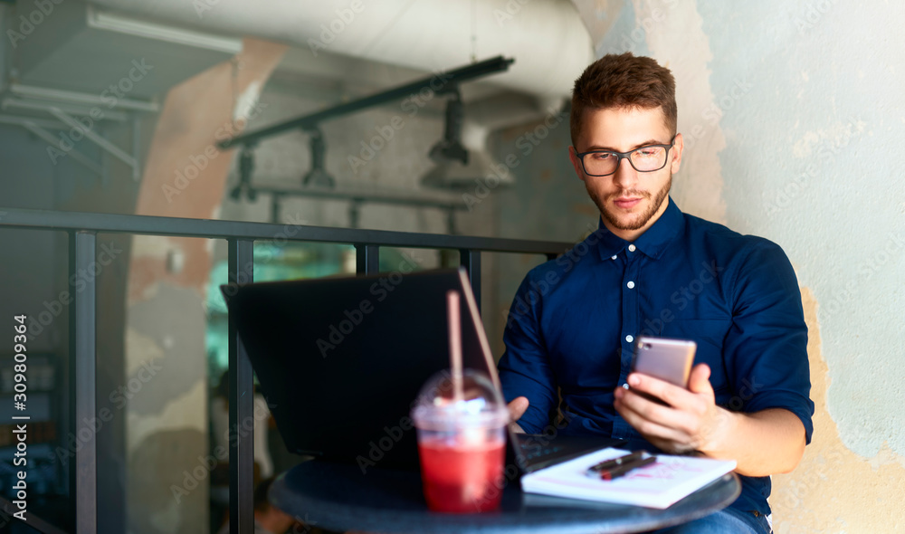 Freelancer typing text on smartphone and working remotely. Hipster holding mobile phone and writing message. Smiling young businessman in glasses telecommuting with cellphone on laptop in loft cafe. Freelancer typing text on smartphone and working remotely. Hipster holding mobile phone and writing message. Smiling young businessman in glasses telecommuting with cellphone on laptop in loft cafe.
