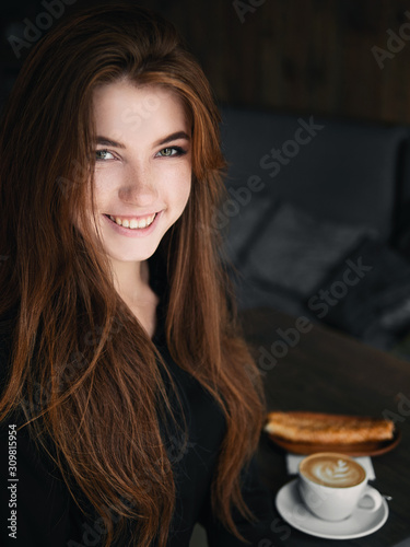 Cute redhead woman woman with adorable smile sitting in cozy city cafe enjoying free time coffee break with cappuccino and dessert