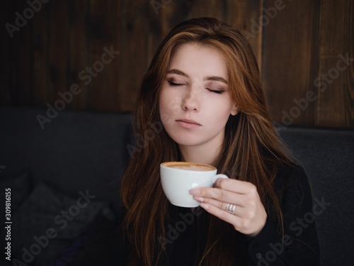 Close up headshot beautiful caucasian red haired woman holding mug drinking cappuccino coffee with latte art adorable smile and feeling good in trendy city cafe