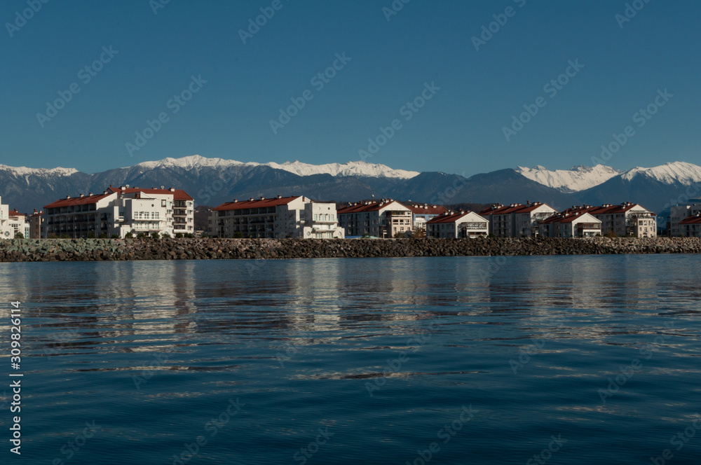 a number of typical four-story houses near the sea. Mountain snow-capped peaks rise above the roofs of houses