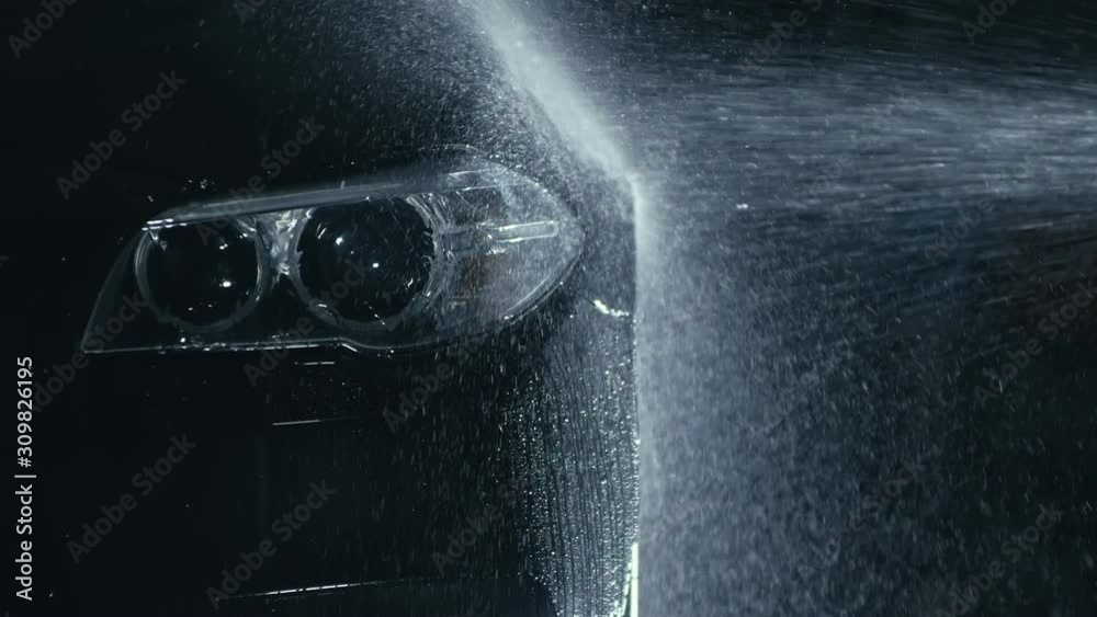 Car headlight wash. Washing modern vehicle body by high pressure jet wash hose water. Auto glass headlamp, angel eyes in drops. Close up of the spotlight of a black automobile as it gets cleaned.
