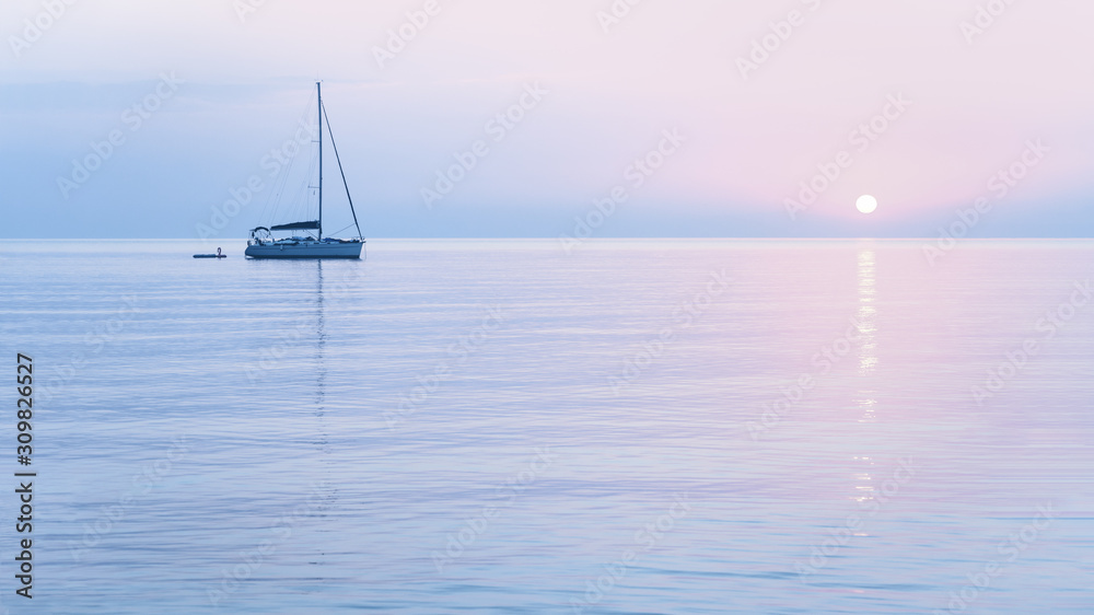 Tender pastel pink sun and sailing boat in blue morning haze color sunrise with blue waters. Travel and vacation destination concept or peace and tranquility concept banner with copy space