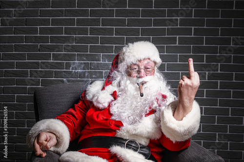 Funny drunk Santa Claus with cigar showing middle finger while sitting in armchair against brick background