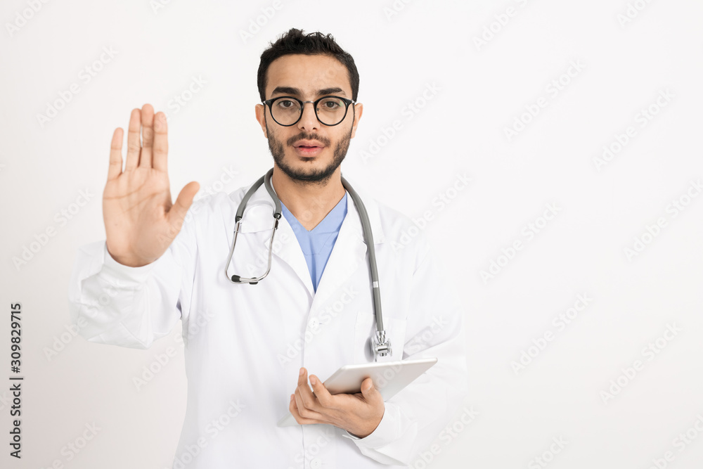 Young practitioner in whitecoat and eyeglasses showing warning sign by hand Young practitioner in whitecoat and eyeglasses showing warning sign by hand